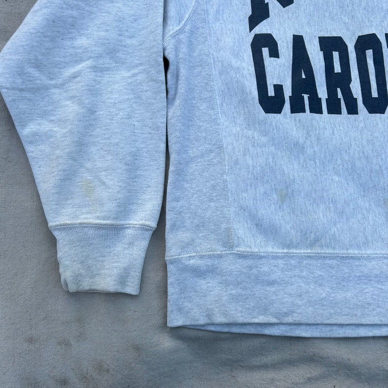 May include: Light grey sweatshirt with the word "CARO" in navy blue block letters. The sweatshirt has a ribbed cuff and hem. The fabric shows some discoloration.