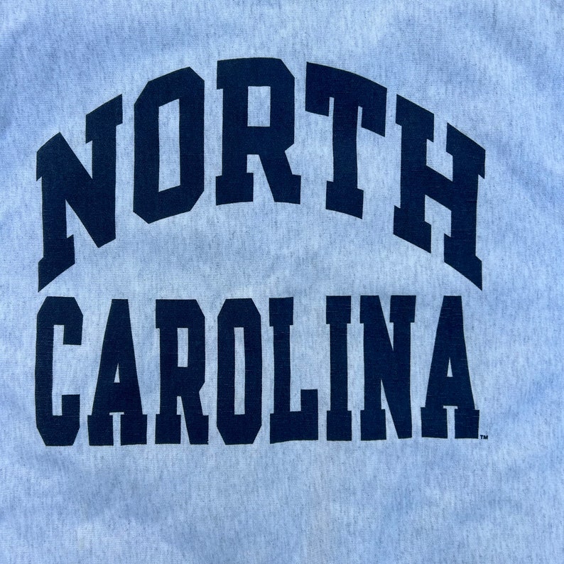 May include: Light grey sweatshirt with the words "NORTH CAROLINA" in navy blue block letters. The text is arched across the top and bottom of the sweatshirt. The fabric has a textured appearance.
