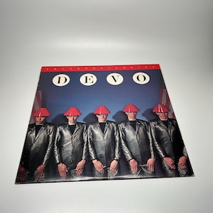 May include: A square vinyl record cover featuring the band Devo. Five individuals in metallic silver jackets and red tiered hats stand against a blue background. The text DEVO appears in white circles, with FREEDOM OF CHOICE in red at the top.