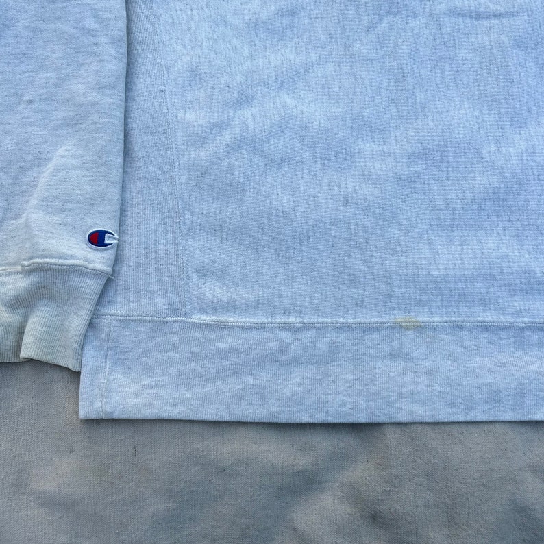 May include: Light gray Champion sweatshirt. The sweatshirt has a crew neck and long sleeves. The Champion logo is visible on the left sleeve cuff. The fabric has a textured appearance.