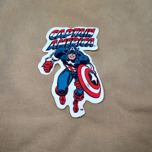 May include: A Captain America patch featuring the superhero in a dynamic pose, holding his shield. The patch has a white border and the words "CAPTAIN AMERICA" in red, white, and blue. The character is in a blue and red costume.