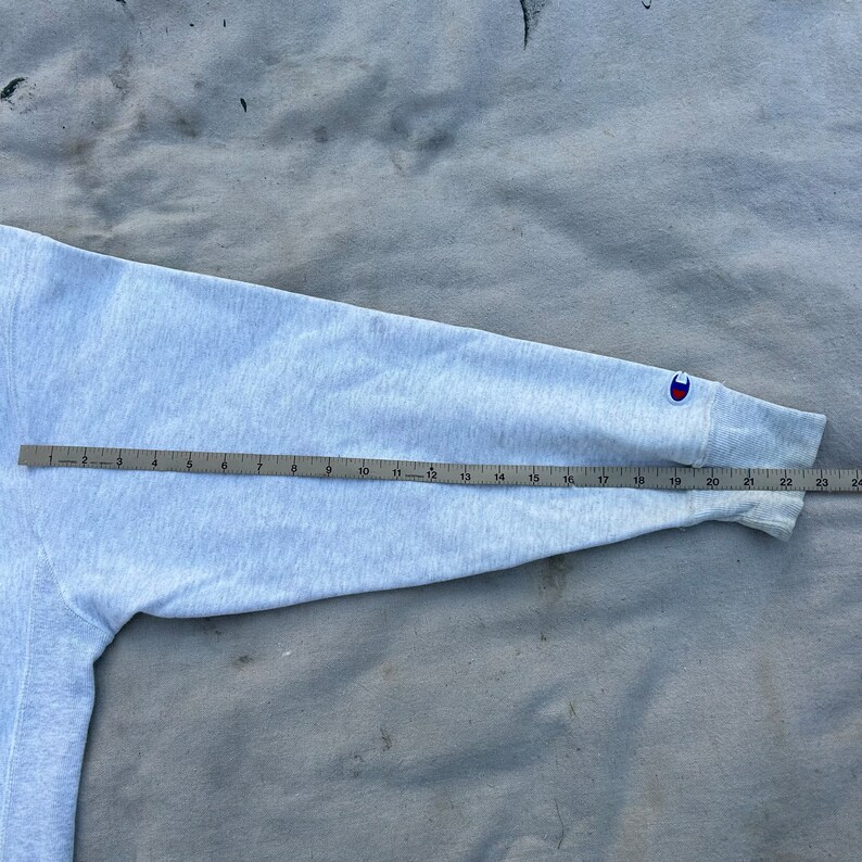 May include: A light gray Champion sweatshirt sleeve with a ruler measuring its length. The sleeve has a small Champion logo on the cuff. The sleeve measures approximately 24 inches in length.