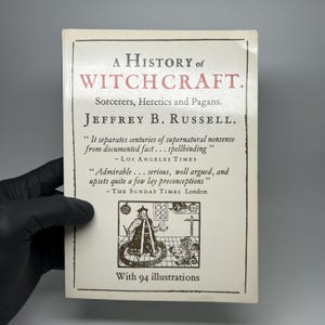 Op de afbeelding: Een boek genaamd "A History of Witchcraft" van Jeffrey B. Russell. De witte omslag heeft zwarte en rode tekst. De titel staat bovenaan, met de naam van de auteur eronder. De omslag bevat citaten en een illustratie.