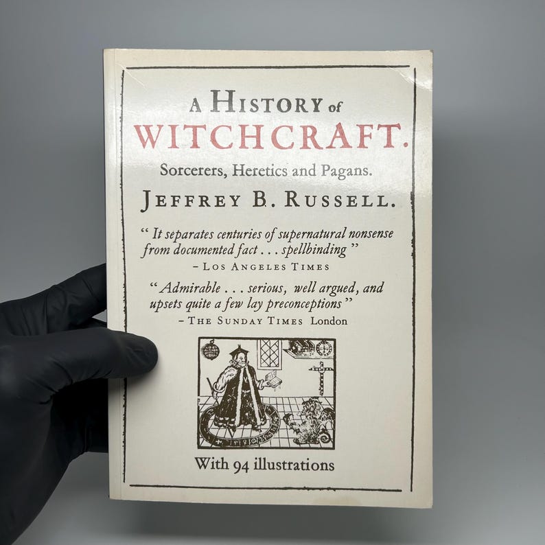 Op de afbeelding: Een boek genaamd "A History of Witchcraft" van Jeffrey B. Russell. De witte omslag heeft zwarte en rode tekst. De titel staat bovenaan, met de naam van de auteur eronder. De omslag bevat citaten en een illustratie.