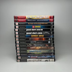 May include: A stack of PlayStation 2 video game cases. Titles include "Sonic Mega Collection Plus", "Kingdom Hearts", "Grand Theft Auto III", "Rayman Arena", and "Scooby-Doo Night of 100 Frights". The cases are in various colors.