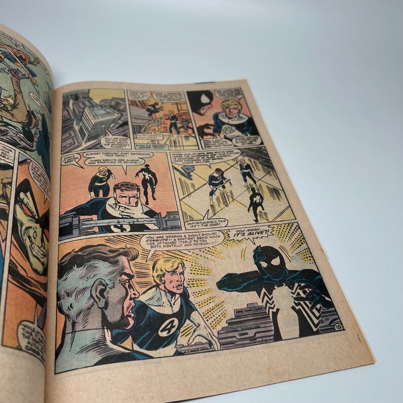 Puede incluir: Una p&aacute;gina de c&oacute;mic vintage con paneles coloridos e ilustraciones de personajes con trajes azules y negros. Los globos de texto incluyen di&aacute;logos como 'You mean... it's alive?!' y 'You are wearing a highly evolved symbiote'.