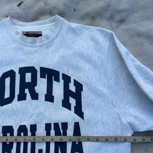 May include: A light gray Champion sweatshirt with the words "NORTH CAROLINA" in navy blue block letters. The sweatshirt has a crew neck and long sleeves. A ruler is placed at the bottom of the sweatshirt.