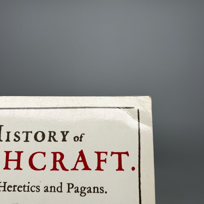 Op de afbeelding: Close-up van een boekomslag met de titel "History of Witchcraft" in zwarte en rode letters. De ondertitel "Heretics and Pagans" is ook zichtbaar. Het boek heeft een witte achtergrond en een grijze achtergrond.