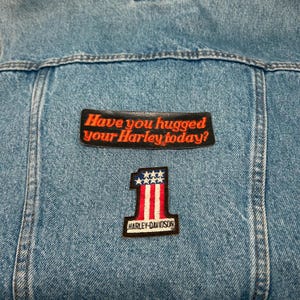 May include: Light blue denim jacket with two patches. The top patch reads "Have you hugged your Harley today?" in orange lettering. The bottom patch is a Harley-Davidson patch with the number "1" and an American flag design.