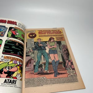 Puede incluir: Un c&oacute;mic abierto que muestra una p&aacute;gina con un anuncio de Mario Bros. de Atari a la izquierda y una vi&ntilde;eta de Spider-Man a la derecha. La vi&ntilde;eta muestra a dos personajes en una habitaci&oacute;n con el texto The Sinister Secret of Spider-Man's New Costume.