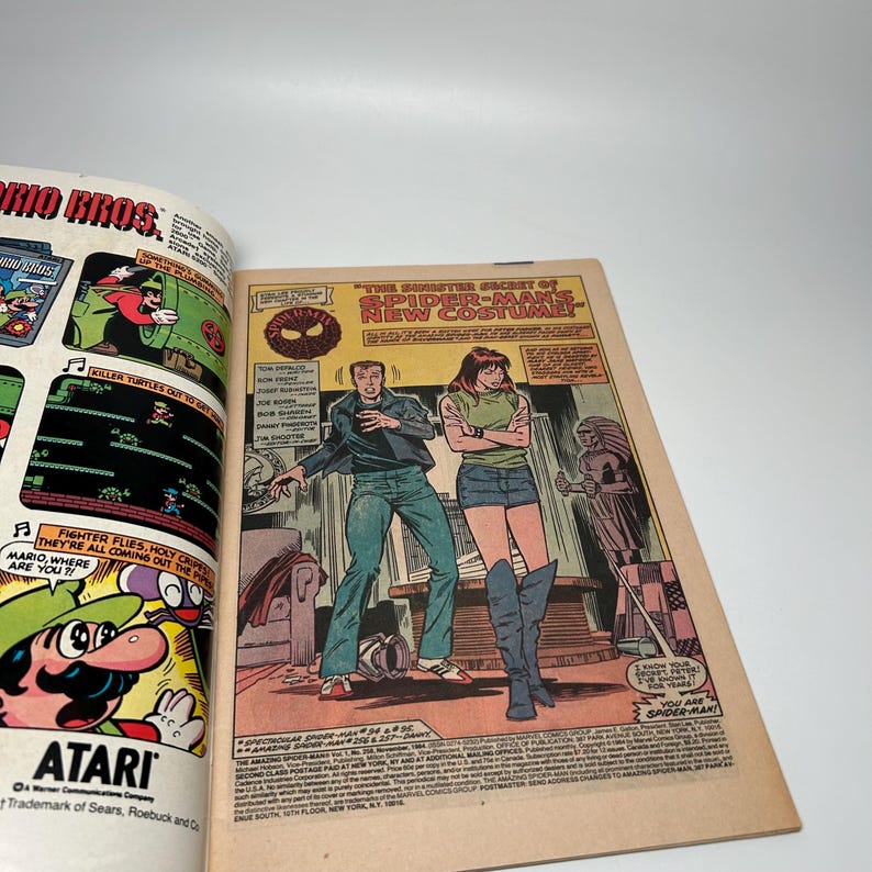 Puede incluir: Un c&oacute;mic abierto que muestra una p&aacute;gina con un anuncio de Mario Bros. de Atari a la izquierda y una vi&ntilde;eta de Spider-Man a la derecha. La vi&ntilde;eta muestra a dos personajes en una habitaci&oacute;n con el texto The Sinister Secret of Spider-Man's New Costume.