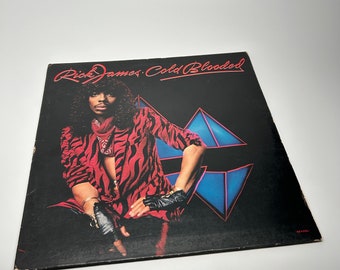May include: A black vinyl record album cover with a photo of a person wearing a red and black tiger print jacket and black gloves. The album title is "Rick James Cold Blooded" in white text.