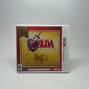 May include: A Nintendo 3DS game case for "The Legend of Zelda: Ocarina of Time 3D." The case features a gold and red design with the game's title and logo. The Nintendo Selects logo is in the upper left corner. The game is rated "E" for Everyone.