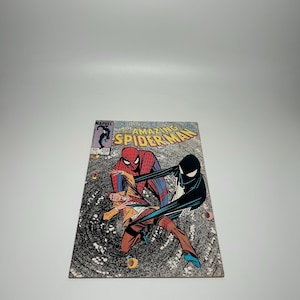 Puede incluir: El n&uacute;mero 258 del c&oacute;mic The Amazing Spider-Man presenta una ilustraci&oacute;n de portada con Spider-Man en su traje cl&aacute;sico rojo y azul, una figura con chaqueta color canela y un personaje de traje negro con un emblema de ara&ntilde;a blanco sobre un v&oacute;rtice.