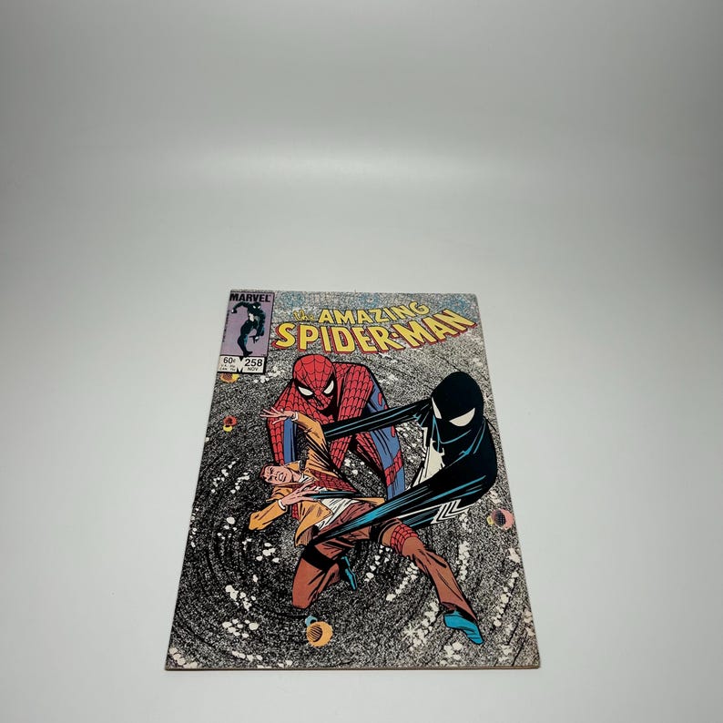 Puede incluir: El n&uacute;mero 258 del c&oacute;mic The Amazing Spider-Man presenta una ilustraci&oacute;n de portada con Spider-Man en su traje cl&aacute;sico rojo y azul, una figura con chaqueta color canela y un personaje de traje negro con un emblema de ara&ntilde;a blanco sobre un v&oacute;rtice.