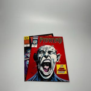 May include: Two comic books are displayed. The front comic book has a red background with a screaming face with pins in the head. The text "PINHEAD" is at the top. The other comic book has a purple and black background.