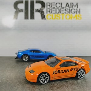 May include: Two die-cast toy cars, one orange and one blue, are positioned on a gray surface. The orange car features the name JORDAN in black lettering on the side with a checkered flag graphic. The text FIR RECLAIM REDESIGN CUSTOMS is visible.