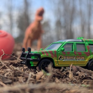 May include: A green and yellow toy SUV with a camouflage pattern and the "Jurassic Park" logo. A red object and a brown dinosaur toy are in the background, set against a blurred outdoor scene.