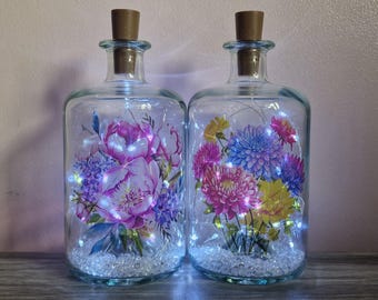 Könnte beinhalten: Zwei Glasflaschen mit Korken, gefüllt mit Lichterketten und Blumenarrangements. Eine zeigt rosa und lila Pfingstrosen, die andere bunte Dahlien. Dekorative Wohnkultur.