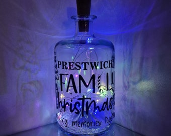 Könnte beinhalten: Eine transparente Glasflasche mit einem schwarzen Korken, gefüllt mit Lichterketten und kleinen, klaren Steinen. Die Flasche trägt den schwarzen Text "PRESTWICH FAMILY Christmas making memories together."