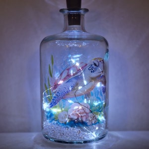 May include: A clear glass bottle with a cork stopper contains a sea turtle illustration with white, blue, and pink accents. The bottle is filled with white fairy lights.