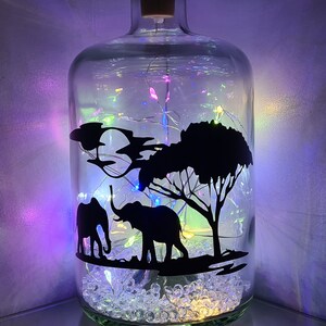 K&ouml;nnte beinhalten: Eine Glasflasche mit einem Korkverschluss, gef&uuml;llt mit klaren Glasperlen und einer Lichterkette. Die Flasche hat eine Silhouette von zwei Elefanten und einem Baum auf der Vorderseite.