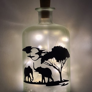 K&ouml;nnte beinhalten: Eine gefrostete Glasflasche mit einem Korkverschluss und einer Silhouette von zwei Elefanten und einem Baum im Inneren. Die Flasche ist mit wei&szlig;en Lichterketten gef&uuml;llt.