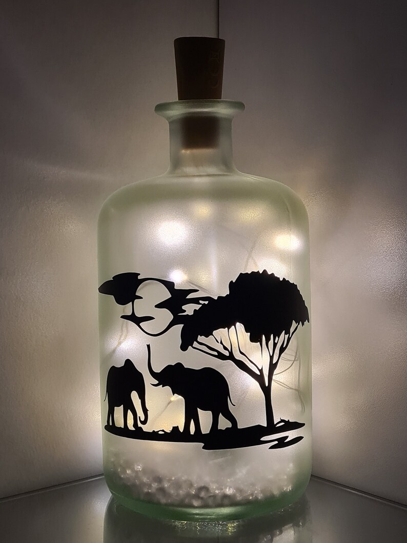 K&ouml;nnte beinhalten: Eine gefrostete Glasflasche mit einem Korkverschluss und einer Silhouette von zwei Elefanten und einem Baum im Inneren. Die Flasche ist mit wei&szlig;en Lichterketten gef&uuml;llt.