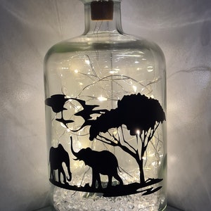 K&ouml;nnte beinhalten: Eine klare Glasflasche mit einem Korkverschluss, gef&uuml;llt mit wei&szlig;en Lichterketten und klaren Glasperlen. Die Flasche hat eine schwarze Silhouette von zwei Elefanten und einem Baum auf der Vorderseite.