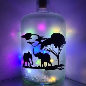 K&ouml;nnte beinhalten: Eine gefrostete Glasflasche mit einer Silhouette von zwei Elefanten und einem Baum im Inneren. Die Flasche ist mit wei&szlig;en Lichterketten gef&uuml;llt, die ein mehrfarbiges Leuchten abgeben.