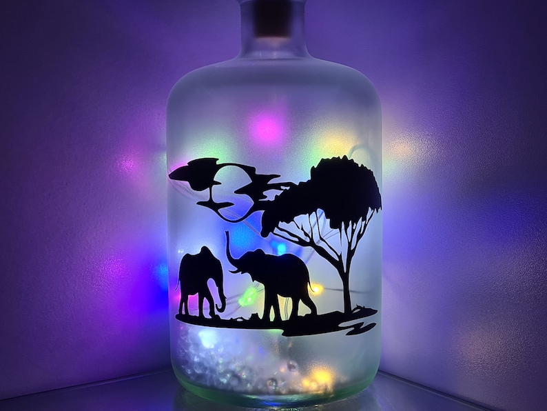 K&ouml;nnte beinhalten: Eine gefrostete Glasflasche mit einer Silhouette von zwei Elefanten und einem Baum im Inneren. Die Flasche ist mit wei&szlig;en Lichterketten gef&uuml;llt, die ein mehrfarbiges Leuchten abgeben.