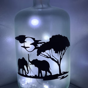 K&ouml;nnte beinhalten: Eine gefrostete Glasflasche mit einem Korkverschluss und einer Silhouette von zwei Elefanten und einem Baum auf der Vorderseite. Die Flasche wird von innen mit wei&szlig;en LED-Lichtern beleuchtet.