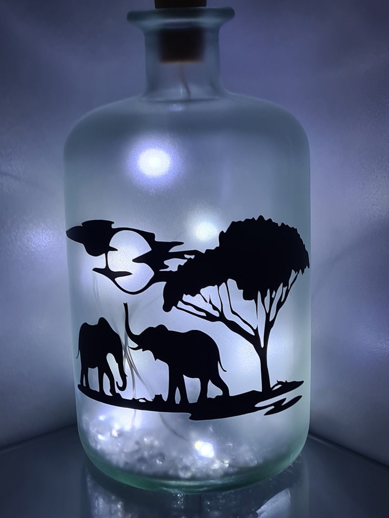 K&ouml;nnte beinhalten: Eine gefrostete Glasflasche mit einem Korkverschluss und einer Silhouette von zwei Elefanten und einem Baum auf der Vorderseite. Die Flasche wird von innen mit wei&szlig;en LED-Lichtern beleuchtet.