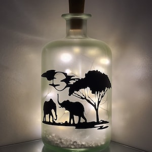 K&ouml;nnte beinhalten: Eine gefrostete Glasflasche mit einem Korkverschluss und einer Silhouette von zwei Elefanten und einem Baum im Inneren. Die Flasche wird von innen mit wei&szlig;en Lichterketten beleuchtet.