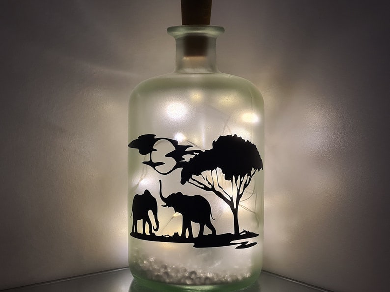 K&ouml;nnte beinhalten: Eine gefrostete Glasflasche mit einem Korkverschluss und einer Silhouette von zwei Elefanten und einem Baum im Inneren. Die Flasche wird von innen mit wei&szlig;en Lichterketten beleuchtet.