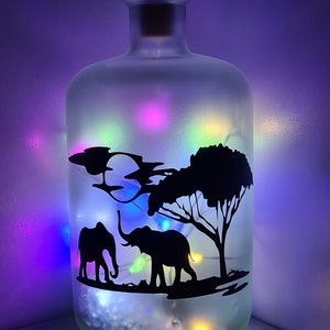 K&ouml;nnte beinhalten: Eine gefrostete Glasflasche mit einem Korkverschluss, verziert mit einer Silhouette von zwei Elefanten und einem Baum. Die Flasche wird mit mehrfarbigen Lichterketten beleuchtet.