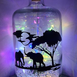 K&ouml;nnte beinhalten: Eine klare Glasflasche mit einem Korkverschluss, gef&uuml;llt mit wei&szlig;en, schimmernden Perlen und einer Lichterkette. Die Flasche zeigt eine schwarze Silhouette von zwei Elefanten und einem Baum.