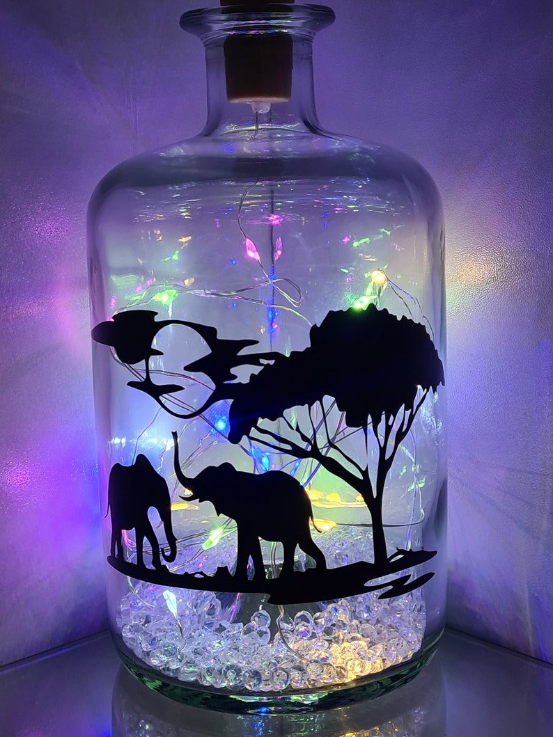 K&ouml;nnte beinhalten: Eine klare Glasflasche mit einem Korkverschluss, gef&uuml;llt mit wei&szlig;en, schimmernden Perlen und einer Lichterkette. Die Flasche zeigt eine schwarze Silhouette von zwei Elefanten und einem Baum.