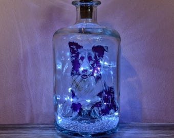 Könnte beinhalten: Eine klare Glasflasche mit Korken, mit einer schwarz-weißen Hundeillustration. Die Flasche wird von kleinen weißen Lichtern beleuchtet und ist mit transparenten Ziersteinen gefüllt. Der Hintergrund ist ein weiches Lila.