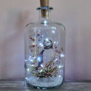 May include: A clear glass bottle with a cork stopper, containing a white bird figurine, white fairy lights, and a floral design. The bottle is sitting on a wooden surface.