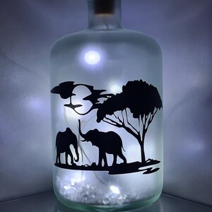 K&ouml;nnte beinhalten: Eine gefrostete Glasflasche mit einer Silhouette von zwei Elefanten und einem Baum. Die Flasche wird von innen mit wei&szlig;en Lichterketten beleuchtet.
