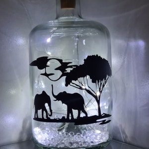 K&ouml;nnte beinhalten: Eine klare Glasflasche mit einem Korkverschluss und einer Silhouette von zwei Elefanten und einem Baum im Inneren. Die Flasche ist mit wei&szlig;en Dekorsteinen und wei&szlig;en Lichterketten gef&uuml;llt.