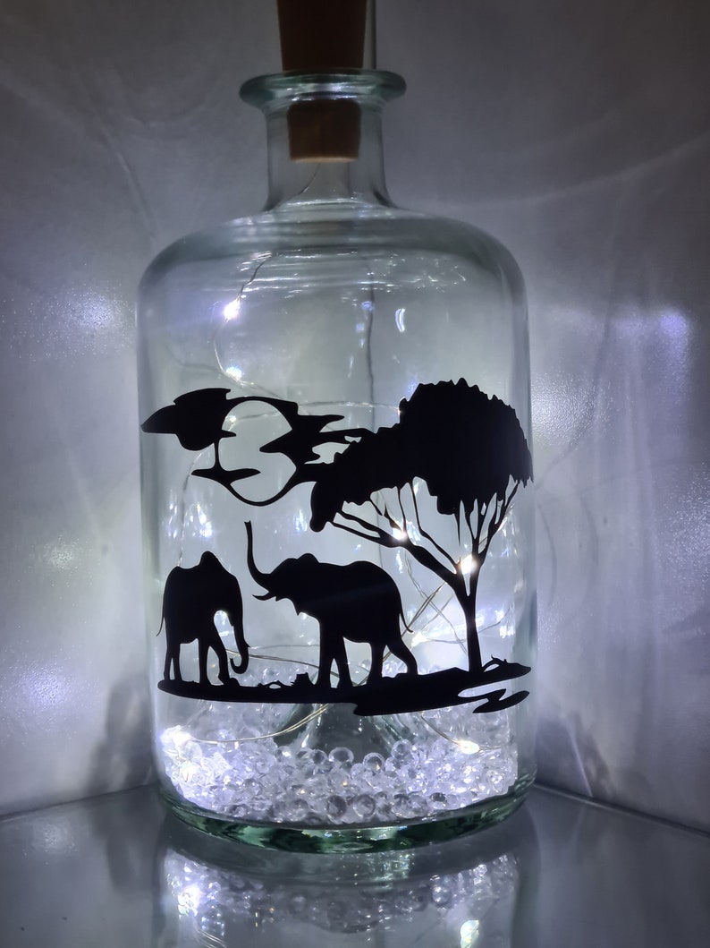 K&ouml;nnte beinhalten: Eine klare Glasflasche mit einem Korkverschluss und einer Silhouette von zwei Elefanten und einem Baum im Inneren. Die Flasche ist mit wei&szlig;en Dekorsteinen und wei&szlig;en Lichterketten gef&uuml;llt.