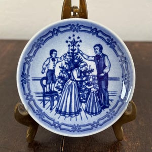 May include: A blue and white porcelain plate featuring a family decorating a Christmas tree. The plate has a detailed border design. The scene depicts a father, mother, and two children. The plate is displayed on a brass stand.