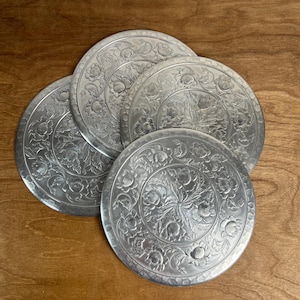May include: A set of three circular silver-toned metal coasters featuring an embossed floral pattern. Each coaster displays a central flower motif surrounded by a ring of smaller blossoms and leaves with a textured, hammered finish.
