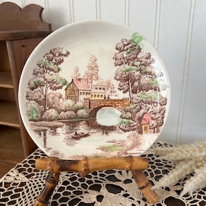 May include: A vintage decorative plate featuring a scenic landscape with a bridge, river, and buildings. The plate has a cream-colored background with brown, green, and tan details. The plate is displayed on a bamboo stand.