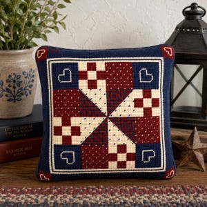 May include: A square needlepoint pillow featuring a geometric pinwheel pattern in navy blue, cream, and deep red. The design includes small heart motifs in the corners and a central star-like arrangement of squares with varied textures and patterns.