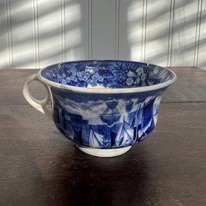 May include: A blue and white ceramic teacup with a handle. The cup features a detailed nautical scene with sailboats and floral patterns. The cup is set on a dark wooden surface.