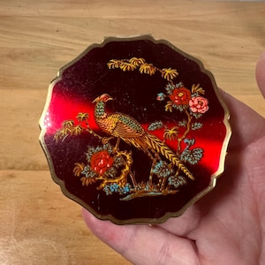 May include: Vintage compact with a red and black background, featuring a detailed illustration of a pheasant perched on a branch with flowers. The compact has a gold-colored trim and a unique, scalloped edge.