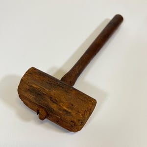 May include: A wooden mallet with a rectangular head and a long, cylindrical handle. The mallet is made of a dark brown wood, with visible grain and texture. The handle is attached to the head at a slight angle.